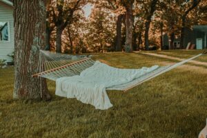 A white hammock with a white blanket draped across it, hanging between two trees in front of a white house.
