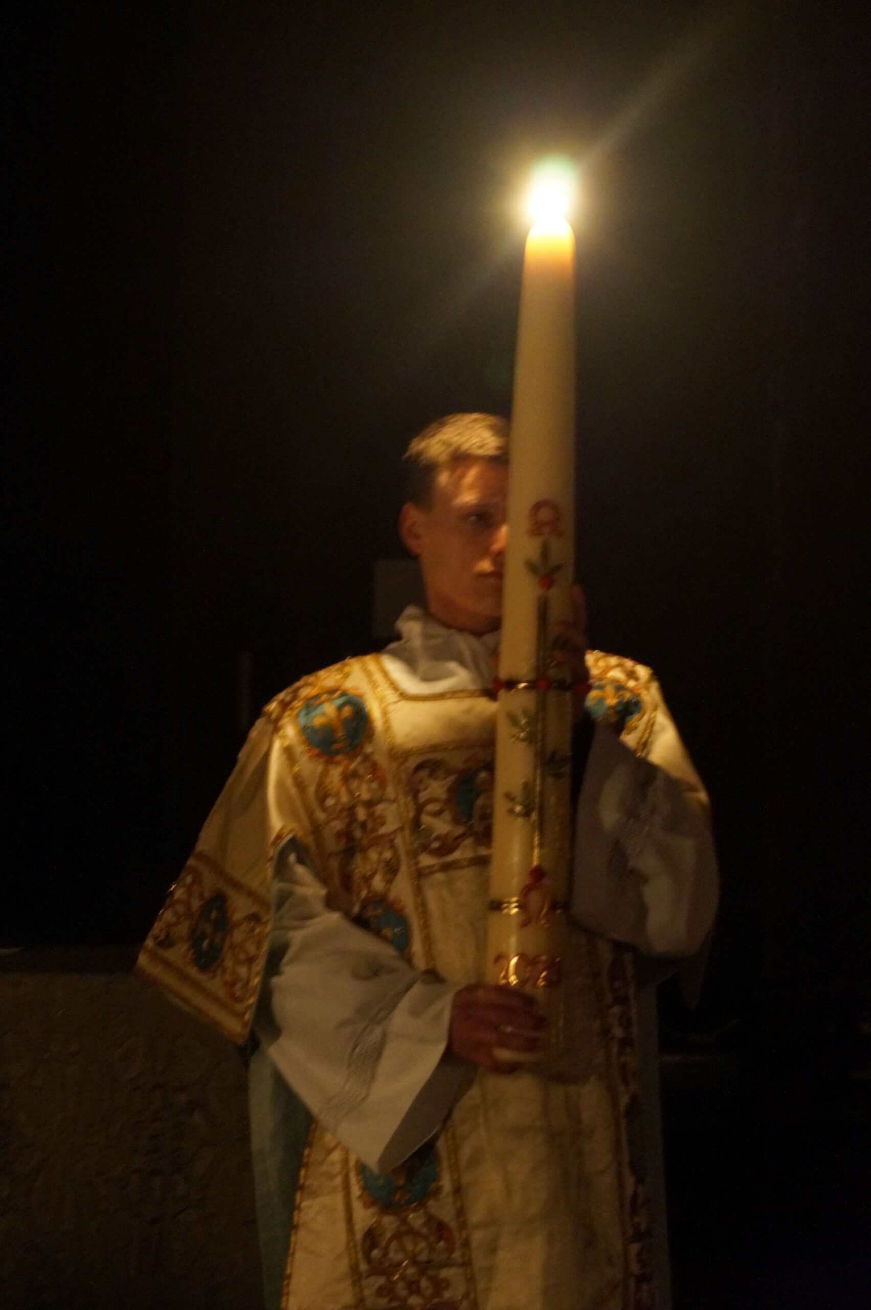 a person in robes, against a dark background, carrying a tall Paschal candle that is lit